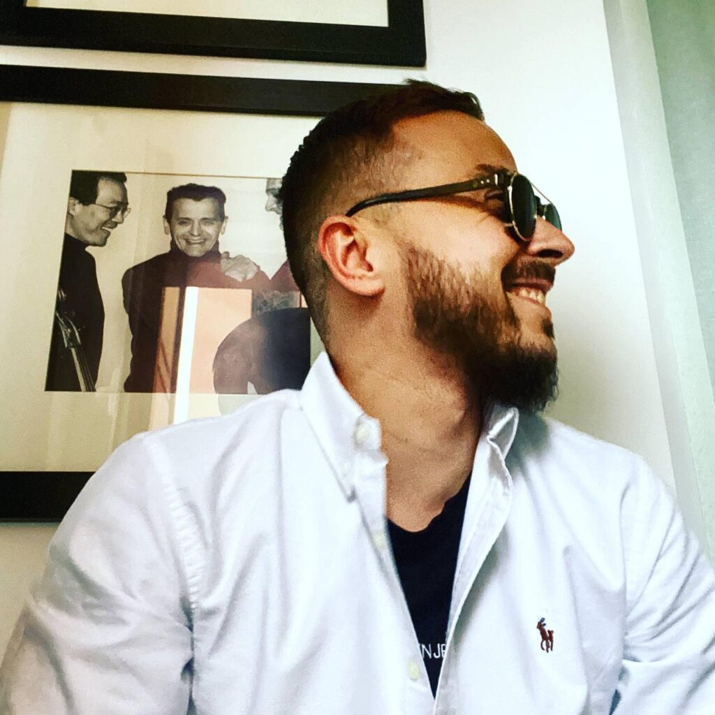 Man in white shirt and sunglasses smiling, with a framed black and white photo in the background.