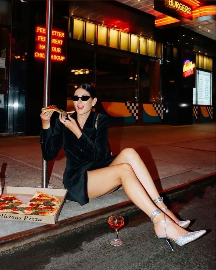 Fashionable woman in sunglasses enjoying a pizza slice on a city street at night, with a box and a cocktail nearby.