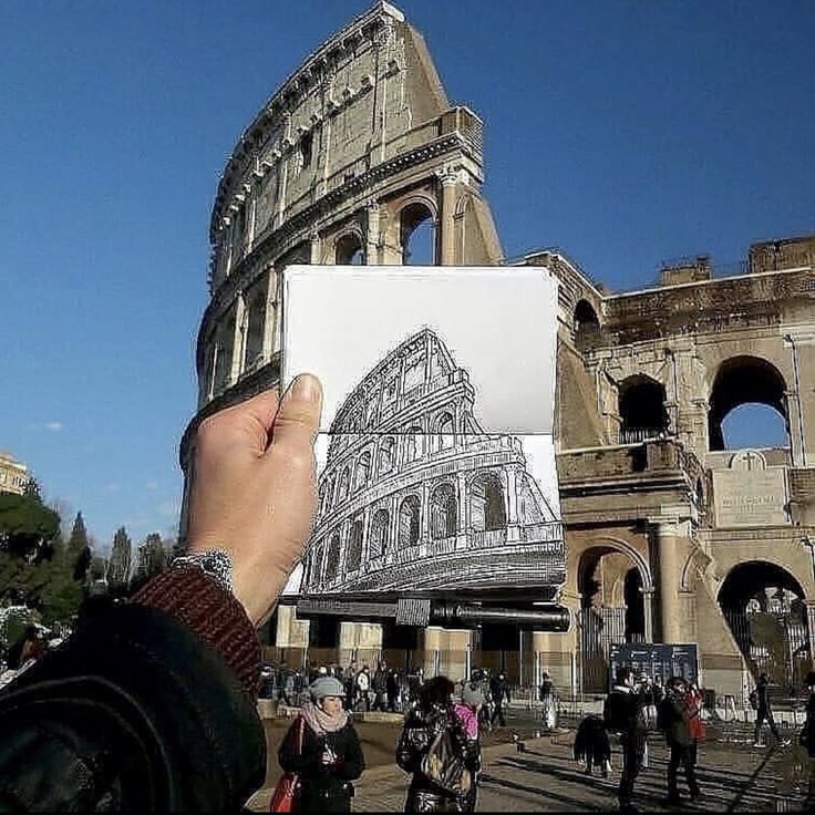 Hand holding a Colosseum sketch aligned with the real structure in Rome, Italy, under a clear blue sky.