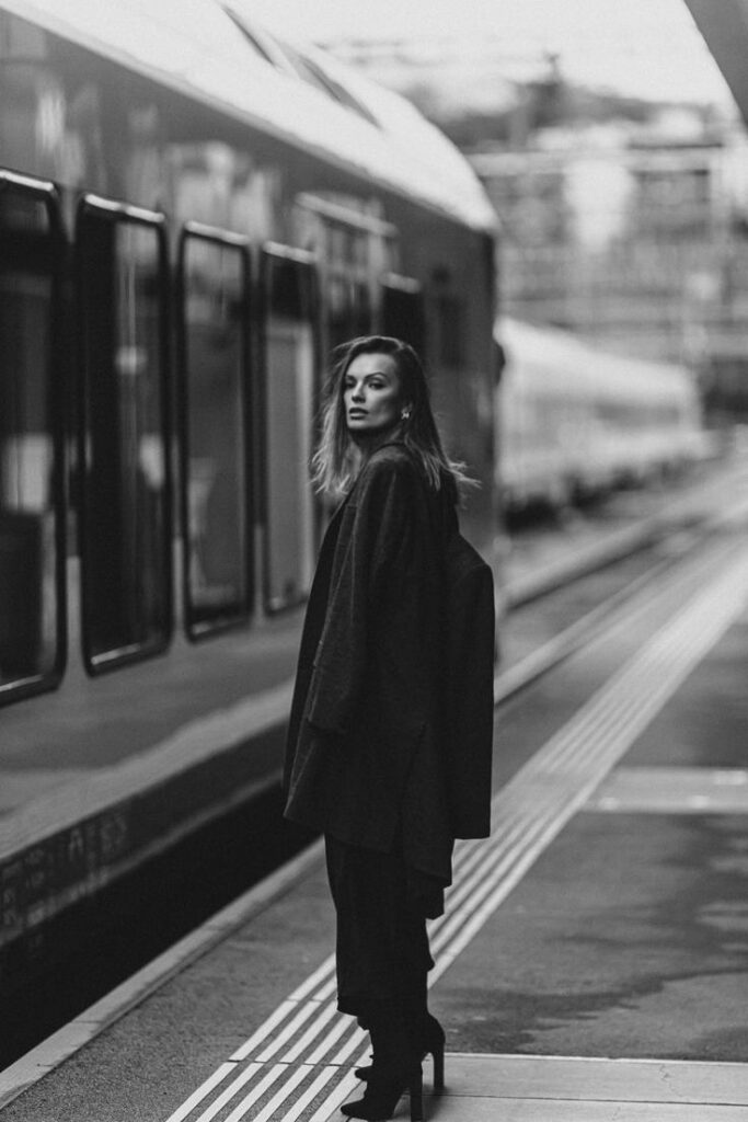 Elegant woman in dark coat walking on train station platform, monochrome style.