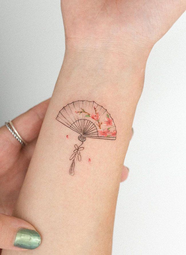 Delicate floral fan tattoo on wrist, featuring pink blossoms and detailed linework, held gently by a hand with green nail polish.