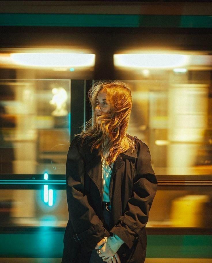 Young woman in a dark coat stands in front of a moving train at night, soft lighting adds a warm glow to the scene.