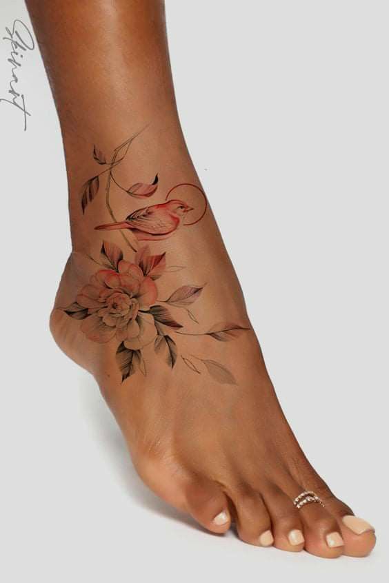 Floral and bird tattoo on ankle with delicate colors and intricate design, adorned with toe rings against a neutral background.