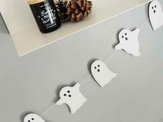 Ghost garland hanging on wall with decorative shelf above, featuring pine cones and a candle. Perfect for Halloween decor.