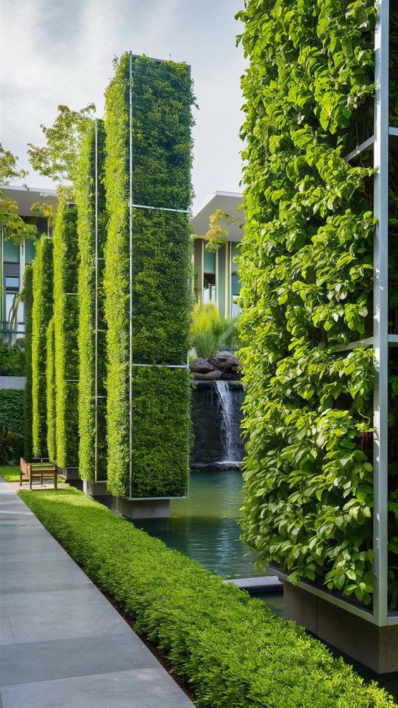 Vertical garden with lush green pillars beside a serene pond and waterfall in a modern outdoor space.