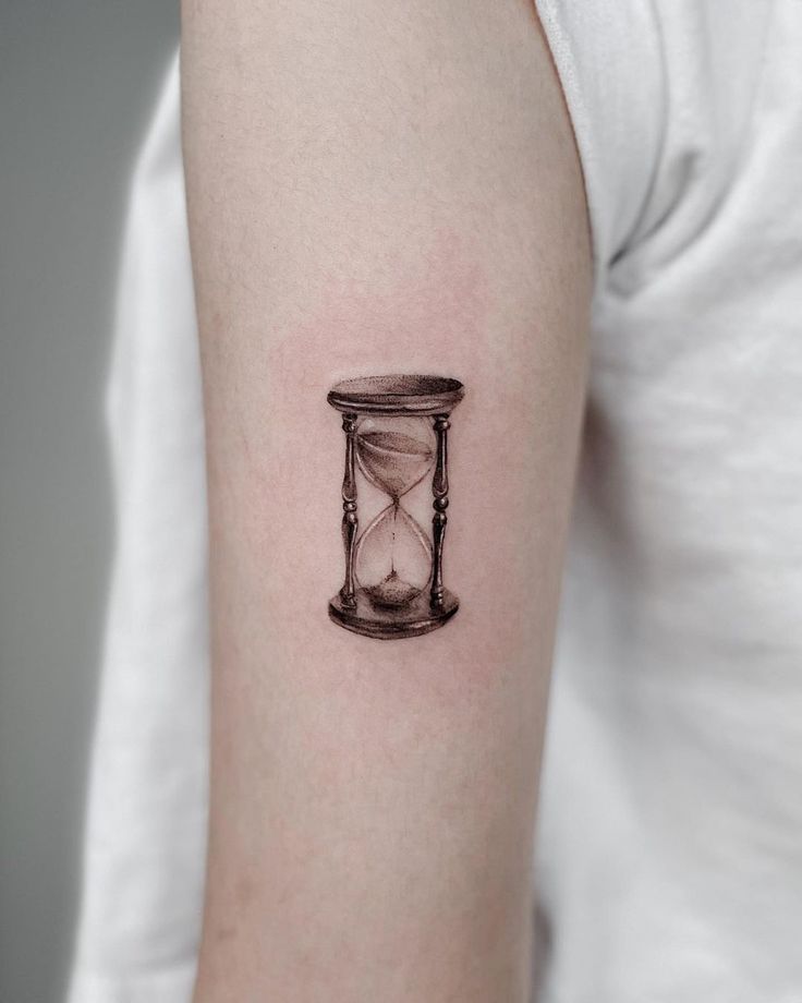 Hourglass tattoo on upper arm, symbolizing time or patience. Simple and detailed black ink design on skin.