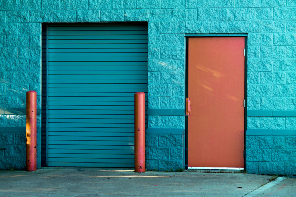 Turquoise garage door and orange entrance on blue brick wall, flanked by bright orange bollards.