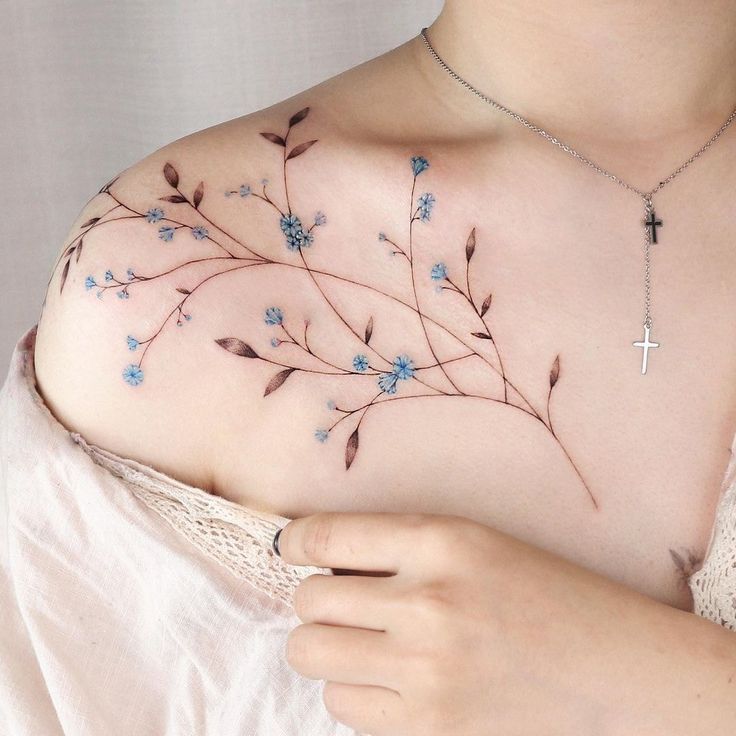 Delicate floral tattoo on shoulder with blue flowers and silver cross necklace.