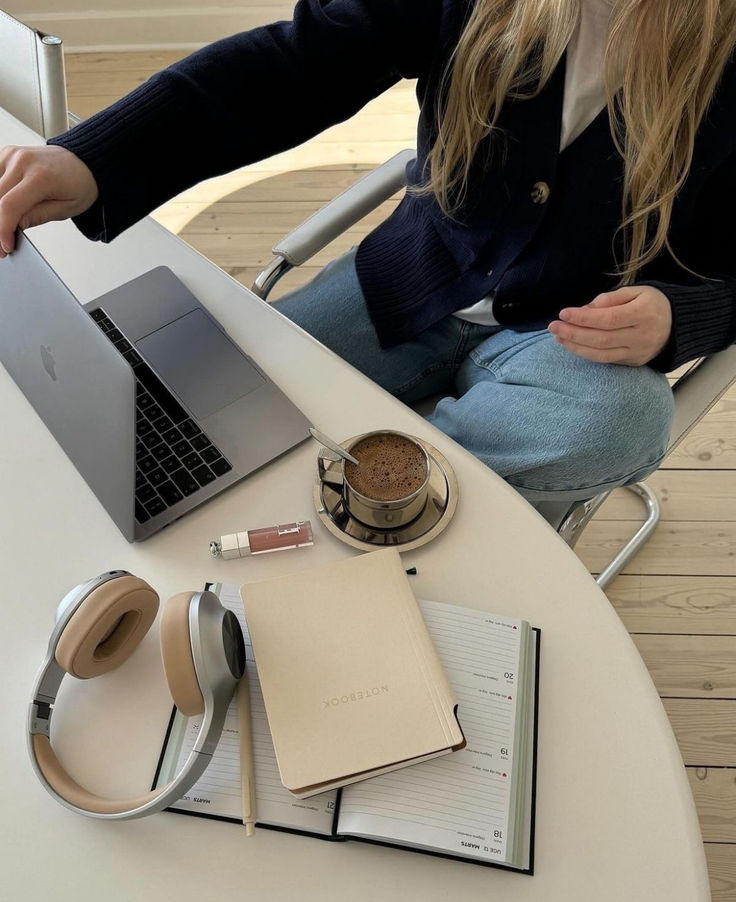 Person working on laptop with coffee, headphones, notebook, and planner on a tidy desk. Cozy workspace setup.