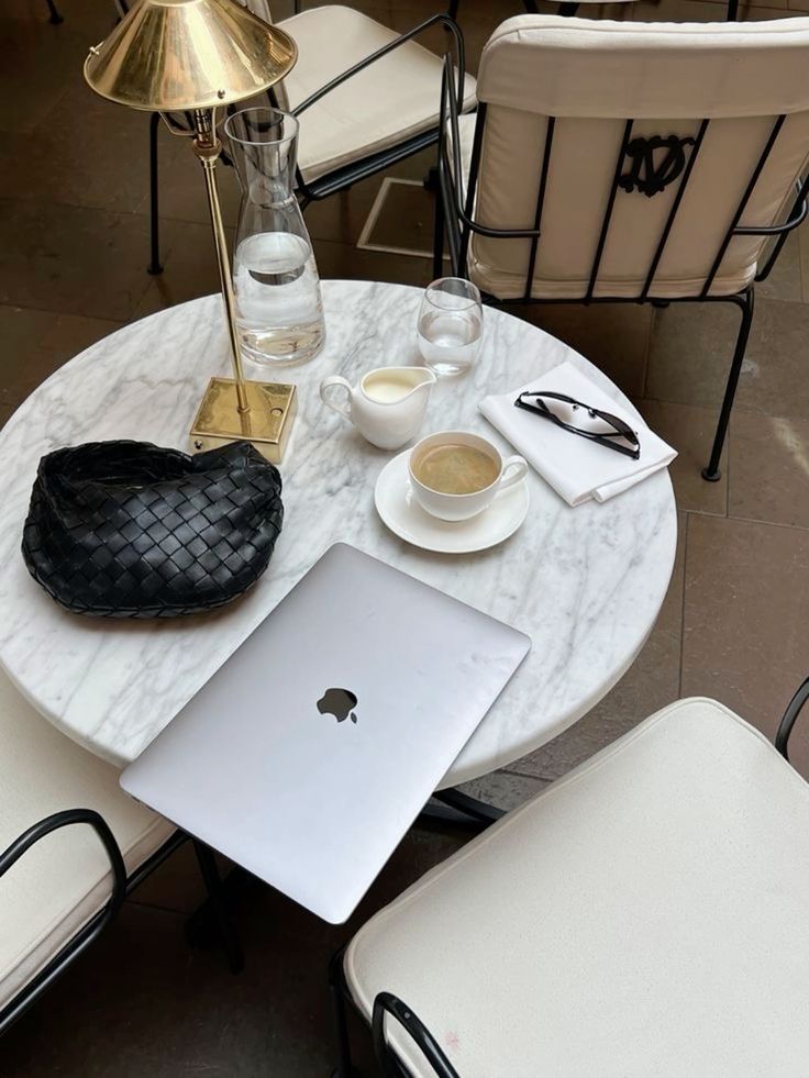 Marble table with laptop, coffee, water carafe, sunglasses, and chic decor in a stylish cafe setting.