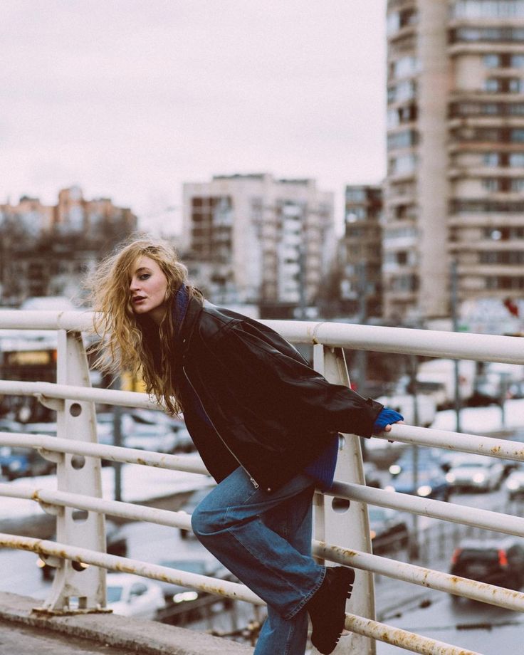 Stylish woman in a black jacket and jeans poses on an urban bridge with city buildings in the background.