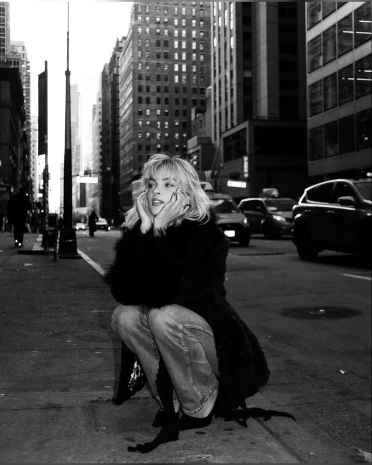 Person crouching on a city street, surrounded by tall buildings and traffic in black-and-white.