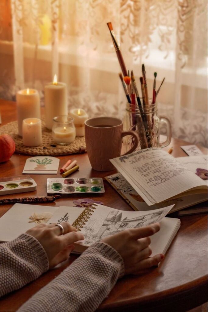 Cozy art scene with candles, sketchbook, paints, and coffee on a wooden table, perfect for creative inspiration.