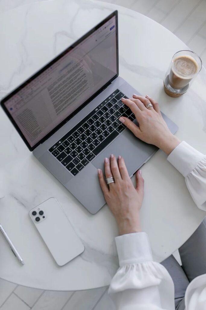 Person typing on a laptop at a marble table with a smartphone and coffee nearby.