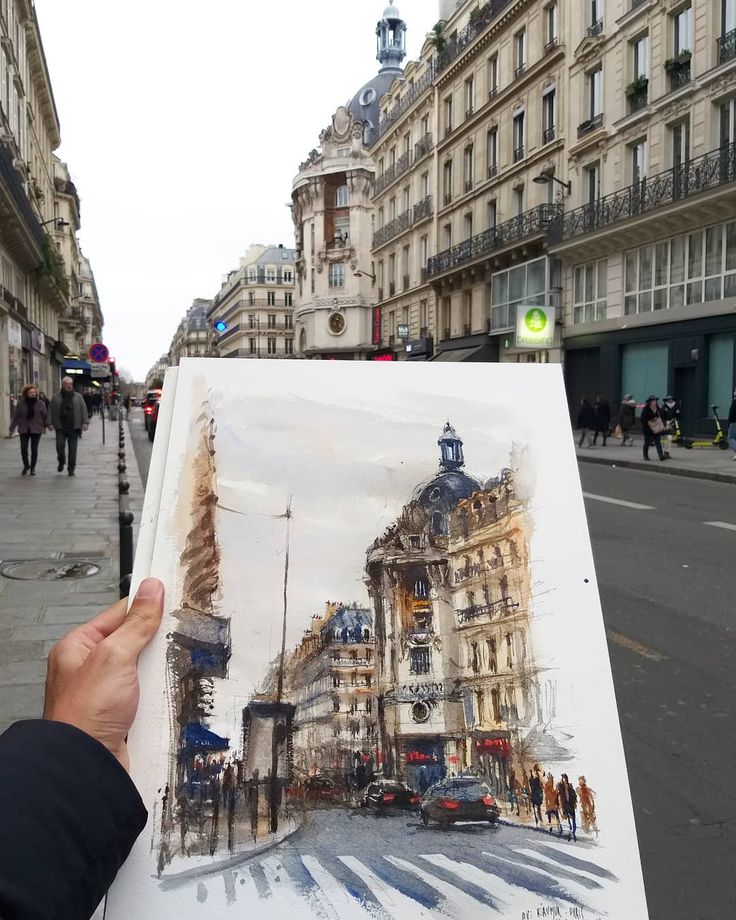 Hand holding painting of a Parisian street scene with ornate buildings and traffic.