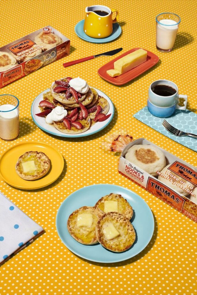 Breakfast table with English muffins, butter, syrup, strawberries, coffee, and milk on a yellow polka-dot tablecloth.