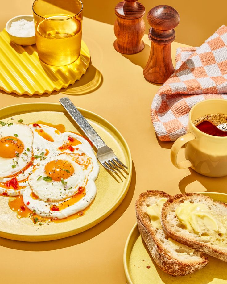 Sunny-side up eggs with herbs, buttered toast, and coffee on a yellow table. Cozy breakfast setting with pepper shakers.