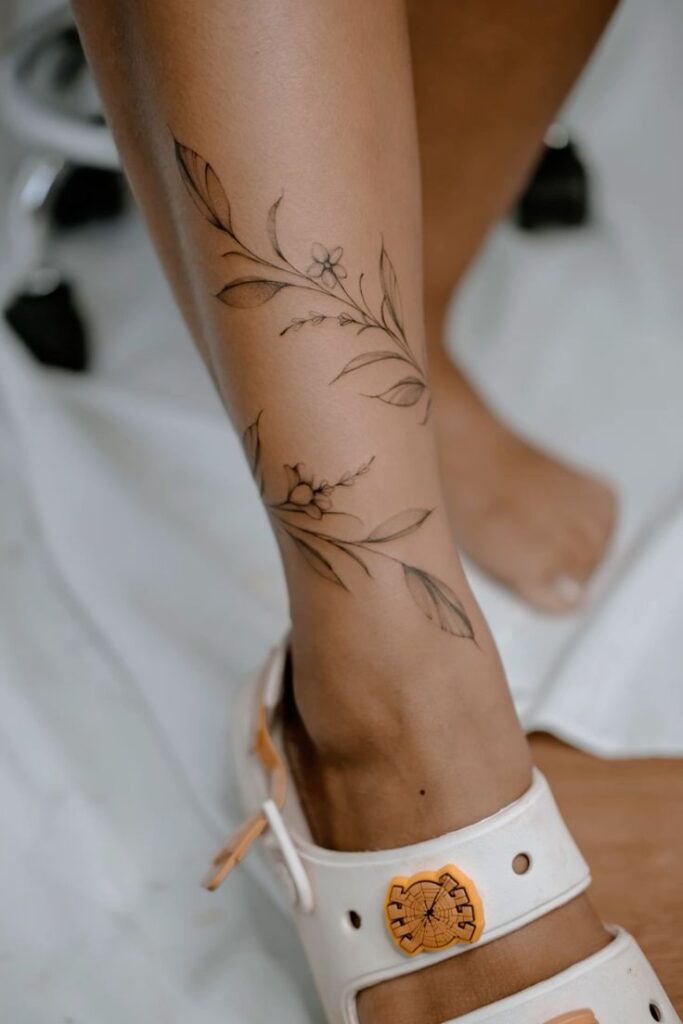 Floral ankle tattoo on woman’s leg, with delicate leaves and blooms, next to a white sandal with an orange charm.