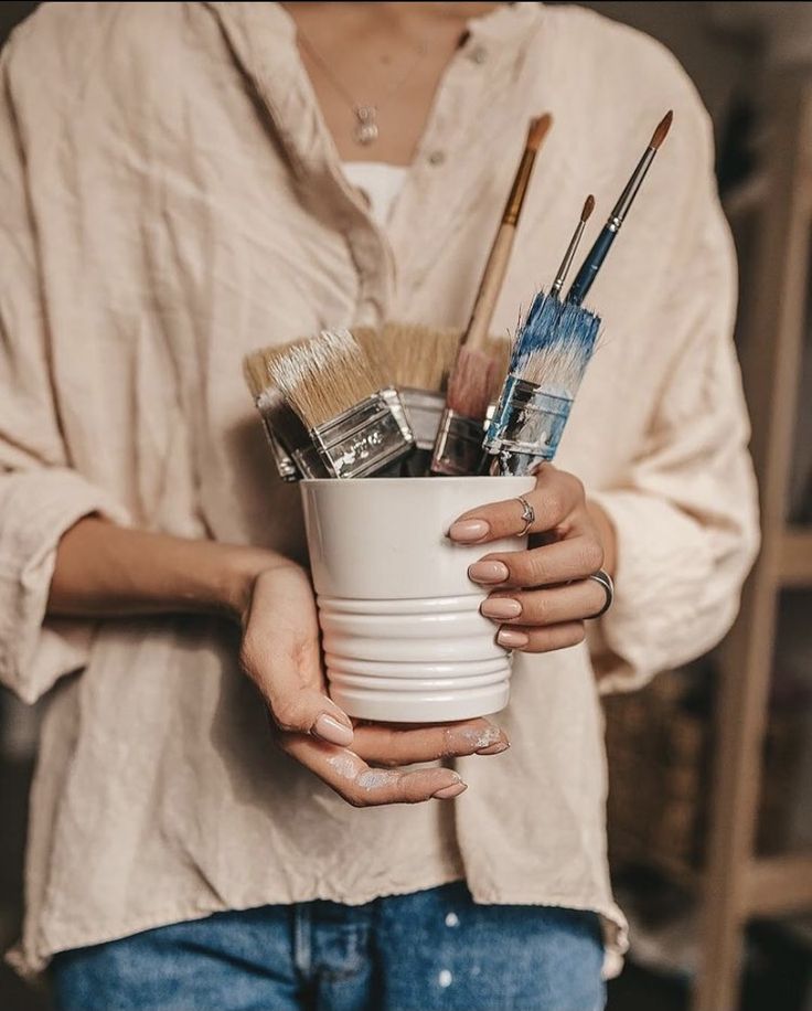 Person holding a white cup filled with various paintbrushes, wearing a beige shirt and blue jeans - creative painting supplies.