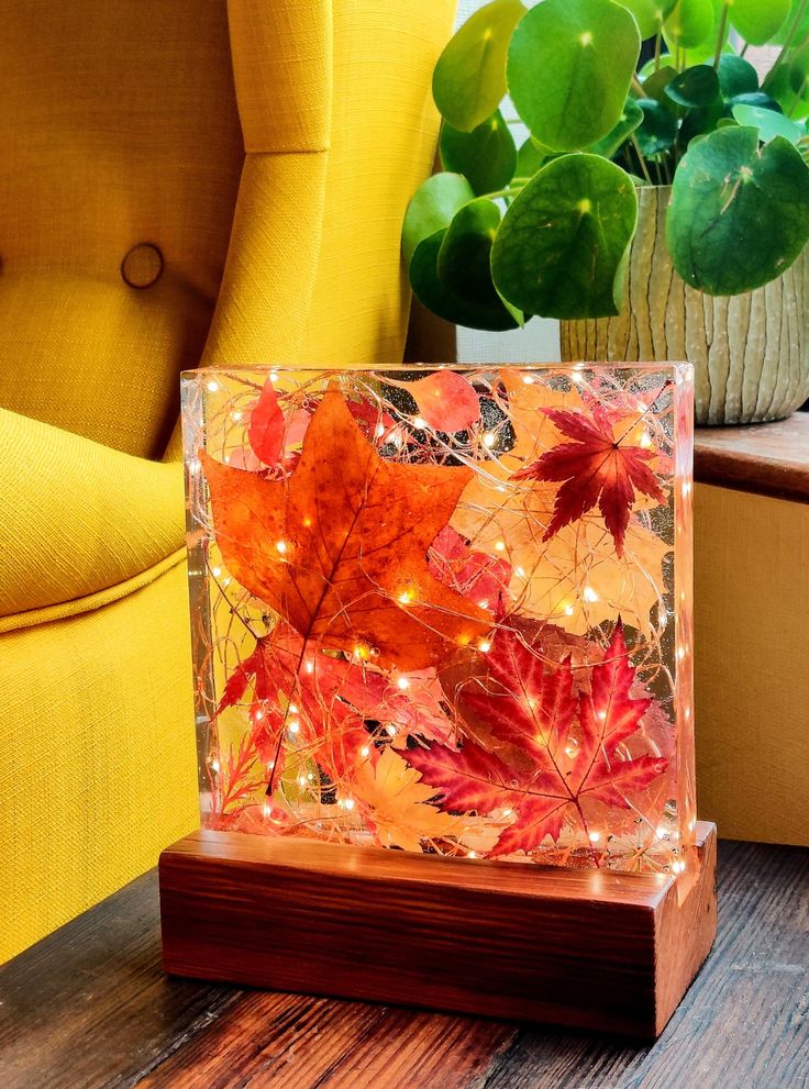 Decorative autumn leaf light box with warm glow, placed by a yellow chair and green plant for cozy home ambiance.