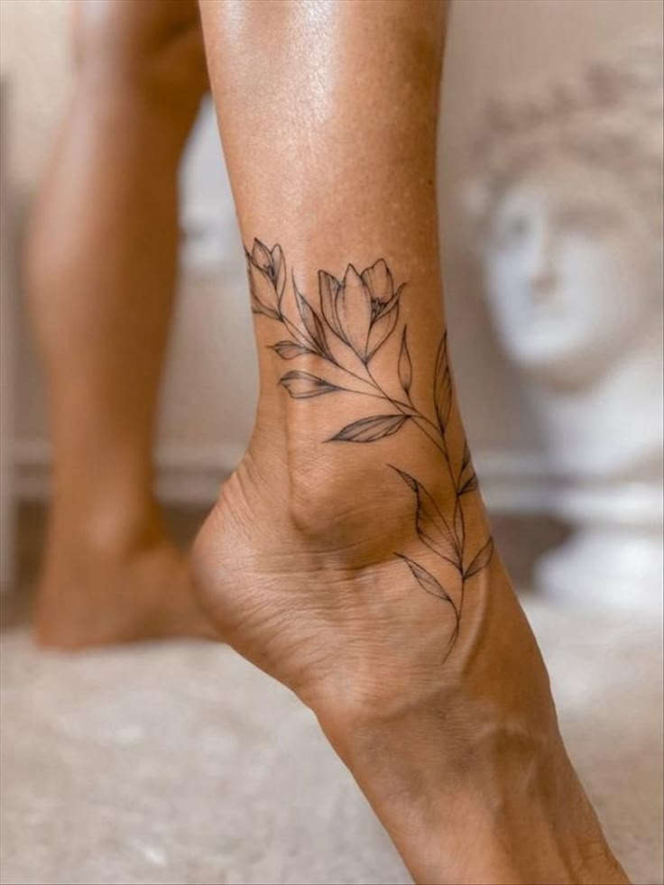 Delicate floral tattoo on ankle, showcasing elegant line work and botanical design against a blurred background.