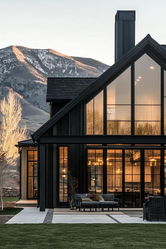 Modern black house with large glass windows, scenic mountain backdrop, and cozy patio at sunset.