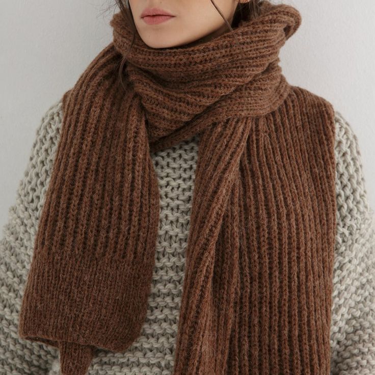 Woman wearing a thick brown knitted scarf and a cozy grey sweater against a neutral background.