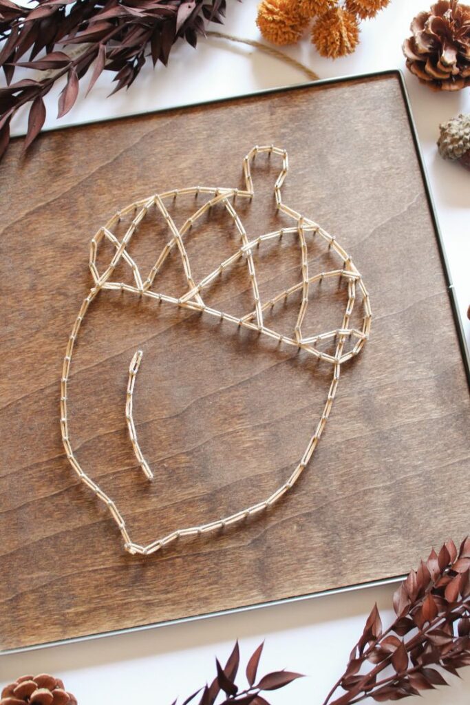 Acorn string art on wood with autumn leaves and pinecones, showcasing a cozy fall decor theme.