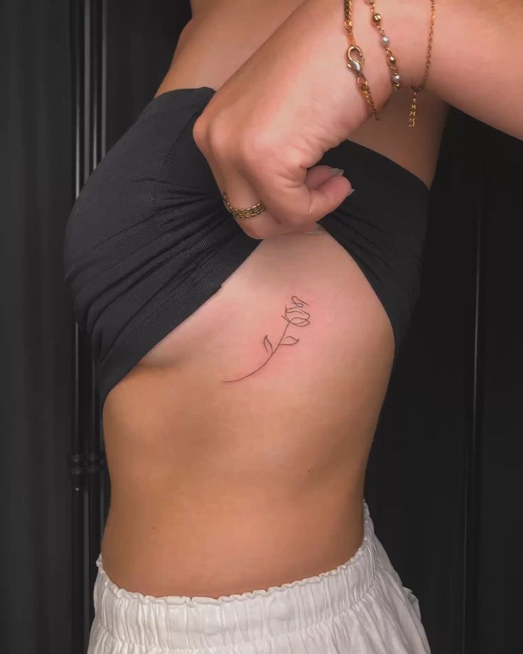 Minimalist flower tattoo on side, woman lifting top to reveal delicate design.