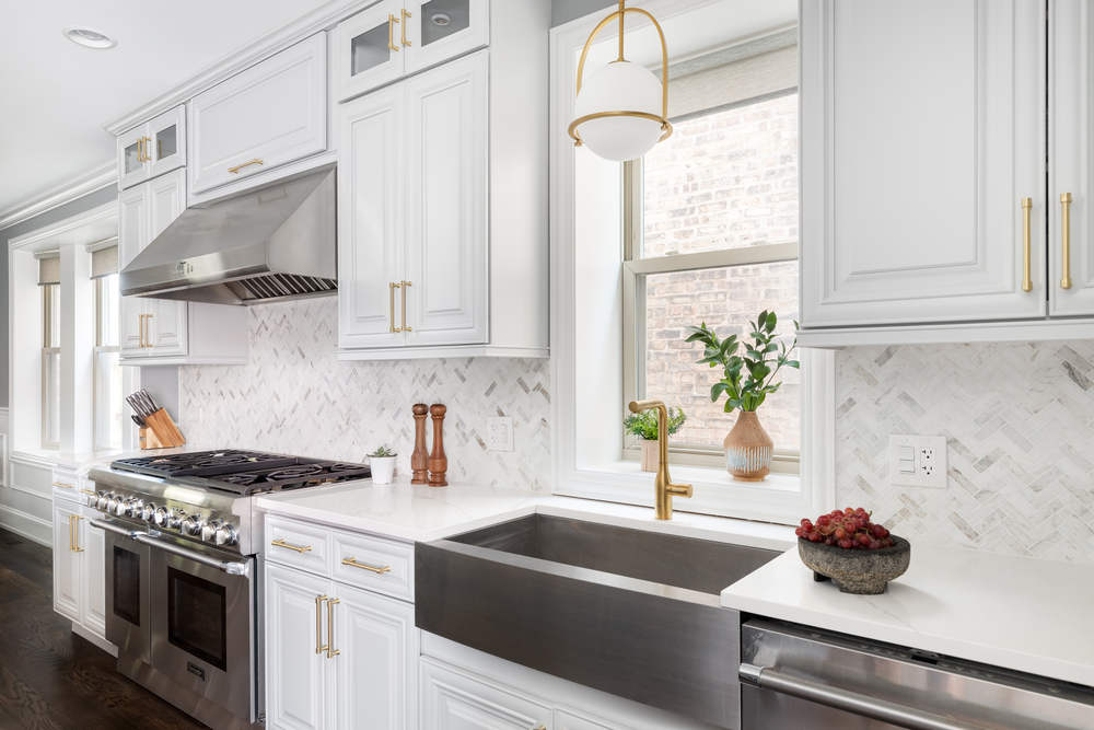 Modern kitchen with stainless steel appliances, farmhouse sink, white cabinets, and gold accents near a sunlit window.