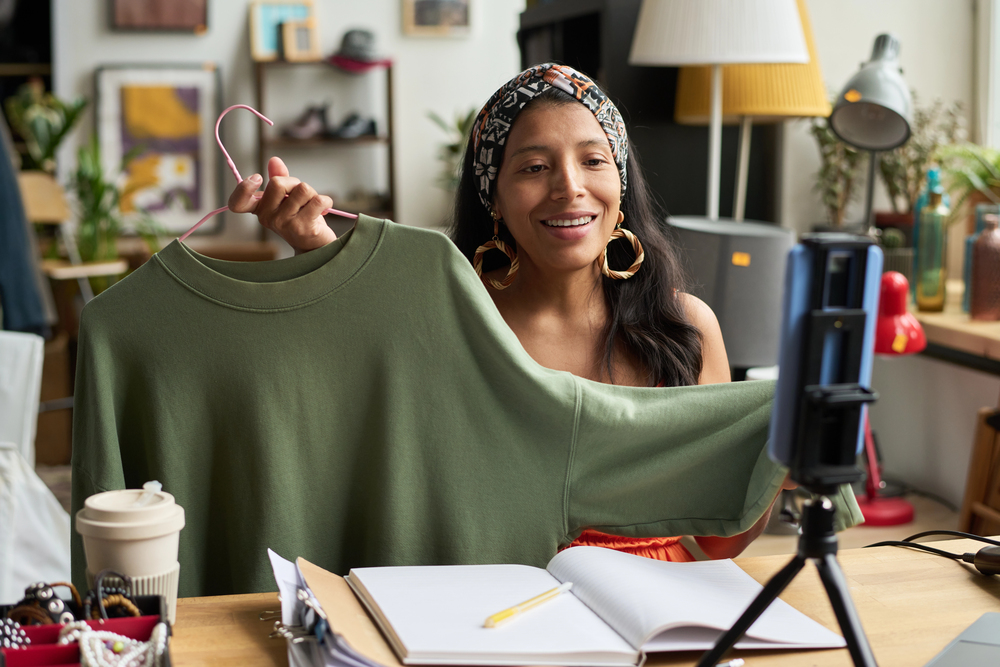 Fashion vlogger showcasing green sweatshirt on video call in cozy home studio.