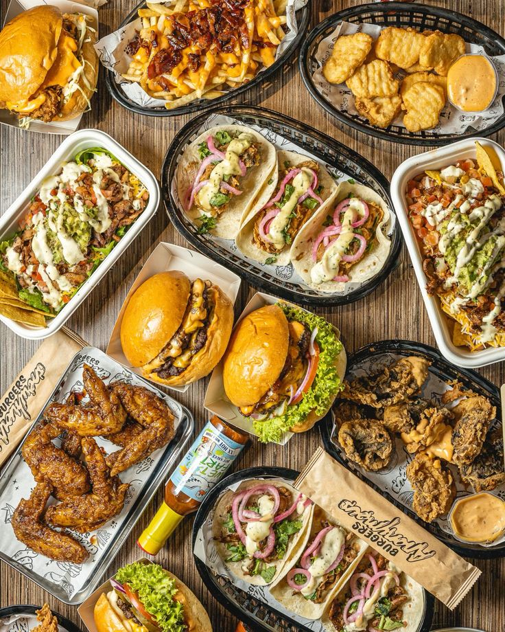 A delicious spread of burgers, tacos, loaded fries, wings, and dipping sauces on a wooden table, perfect for a feast.