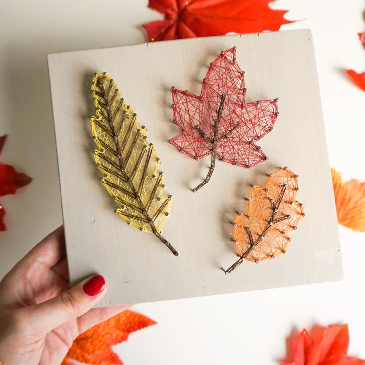 String art featuring colorful autumn leaves on a wooden board, showcasing red, yellow, and orange hues. Perfect for fall decor.