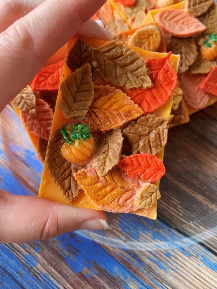 Hand holding a fall-themed soap with colorful leaves and small pumpkins on a wooden background.