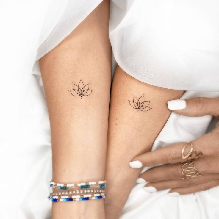 Matching minimalist lotus tattoos on two wrists, adorned with bracelets and rings, close-up.