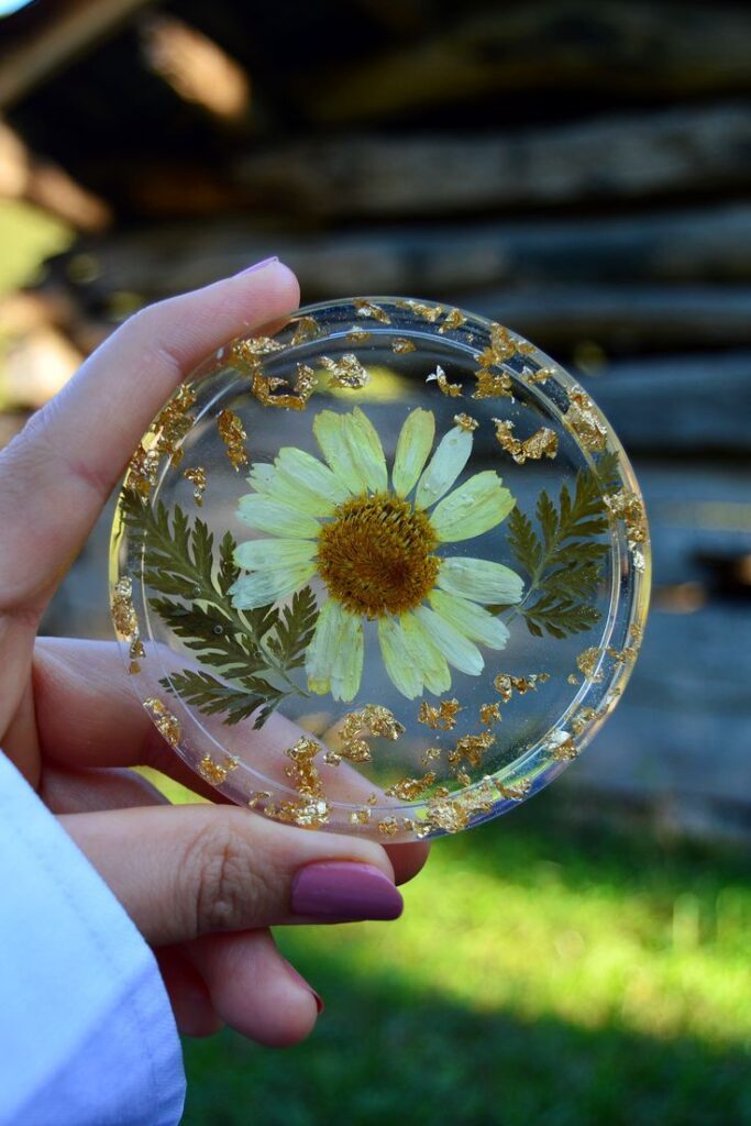 Hand holding floral resin art with pressed daisy and gold flakes, rustic background. Perfect for nature-inspired decor.