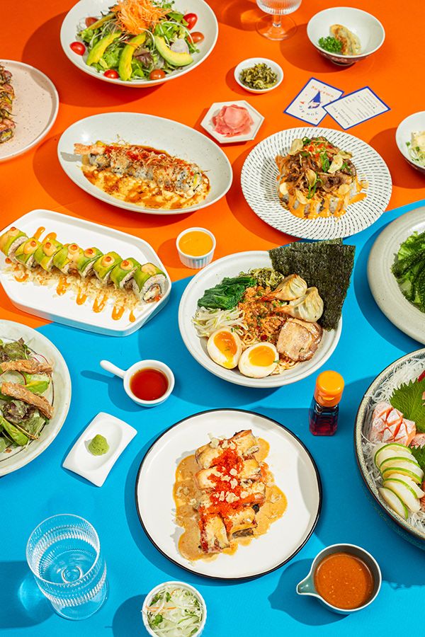 Colorful Asian cuisine assortment with sushi, ramen, and salads on vibrant orange and blue mats.