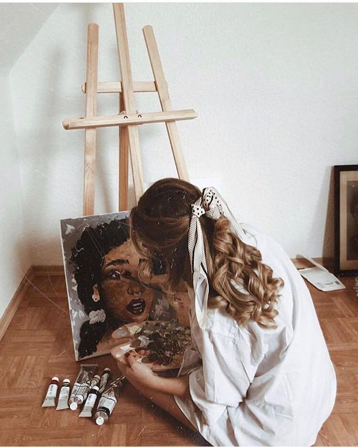 Artist painting a portrait on canvas, sitting on the floor next to an easel, with paint tubes and brushes nearby.