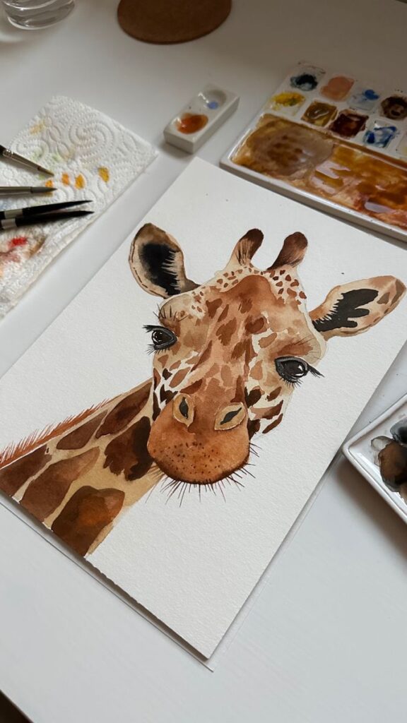 Watercolor painting of a giraffe surrounded by brushes and a palette on a white table.