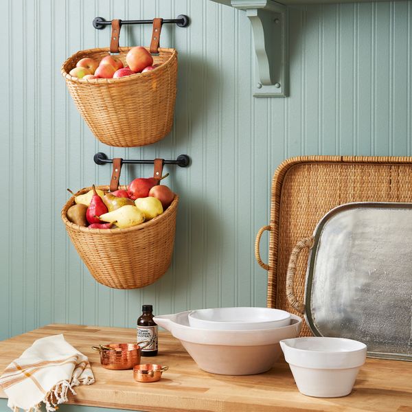 Rustic kitchen with wicker baskets of fruit on wall, wooden counter with bowls, trays, copper measuring cups, and bottle.