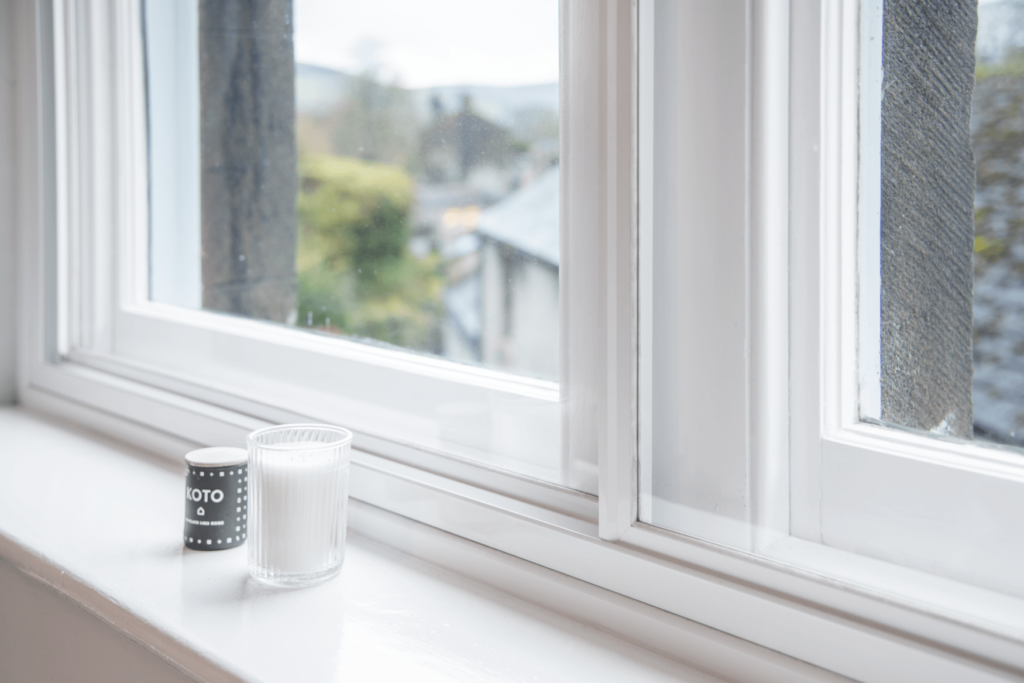 White windowsill with a black and white Koto candle beside a glass window, overlooking a blurred outdoor view.