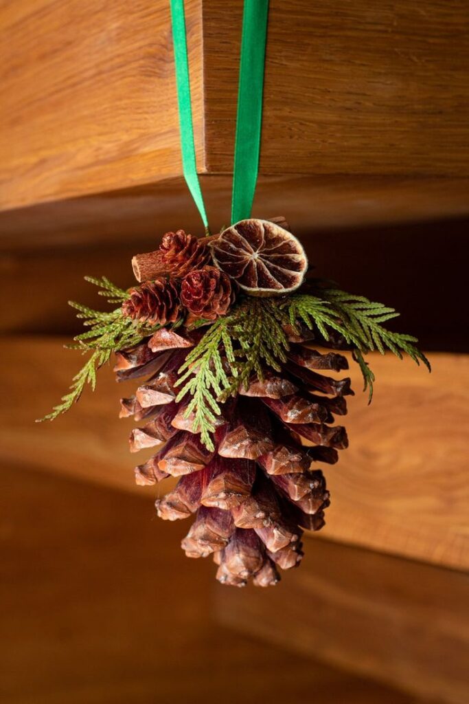 Festive pine cone decoration with sprigs and dried orange, hanging by a green ribbon on wooden background.