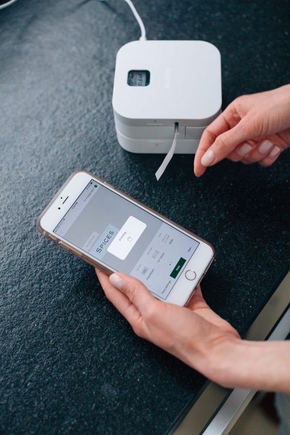 Person using a smartphone to print a SPICES label wirelessly via a compact label printer on a black surface.