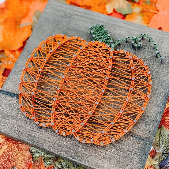 Pumpkin string art on wood with autumn leaves background, perfect for fall decor or craft inspiration.