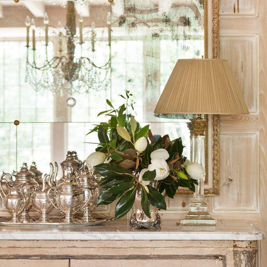 Elegant silver tea set and floral arrangement on vintage dresser with lamp and ornate mirror in luxurious room.