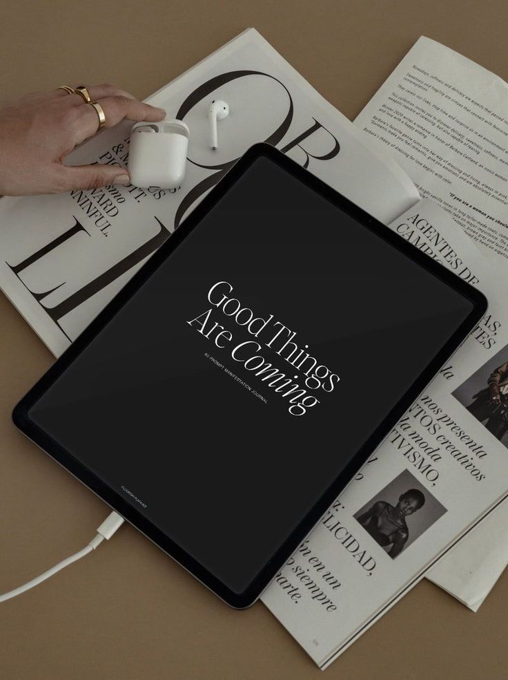 Tablet displaying Good Things Are Coming message beside magazines, AirPods in a case, and hand on table.