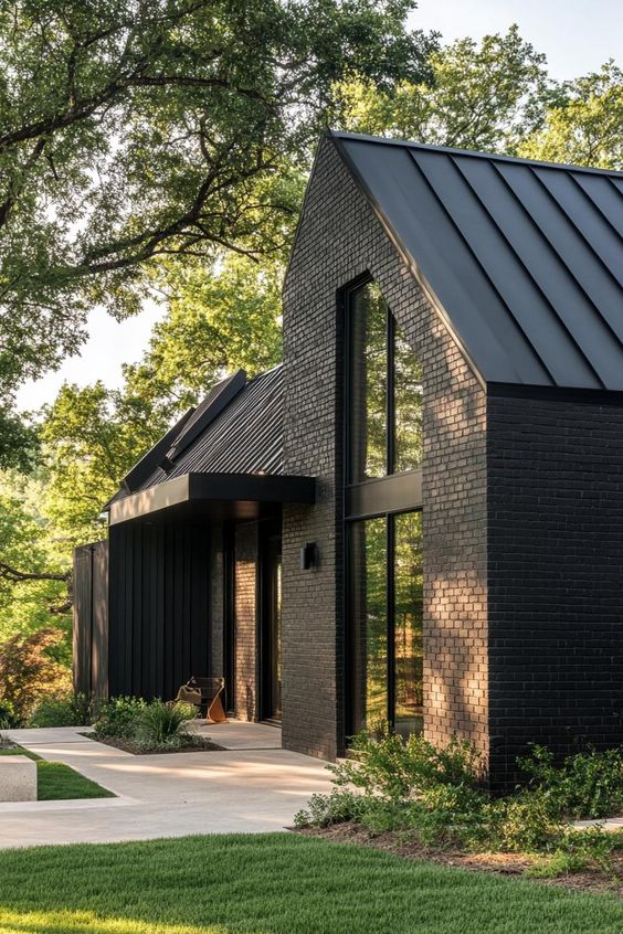 Modern black brick house with large windows, surrounded by lush greenery and trees, with a paved walkway in front.