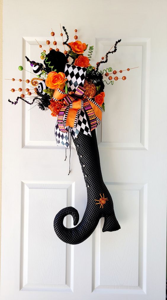 Halloween wreath with orange flowers and ribbons in a witch boot shape, hanging on a white door.