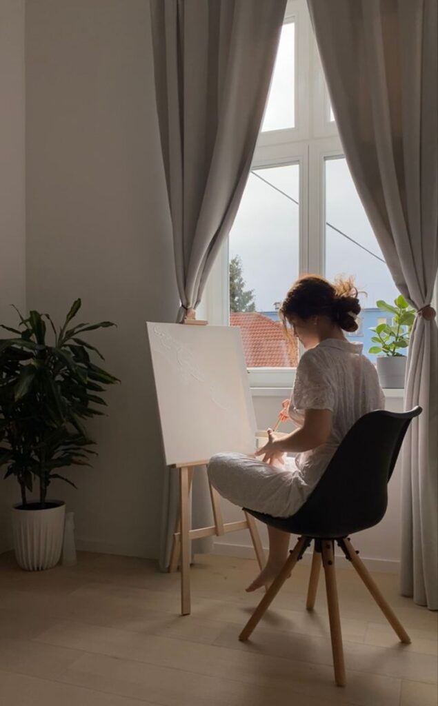 Woman painting on a canvas in a bright room with large windows, plants, and curtain, while sitting on a black chair.