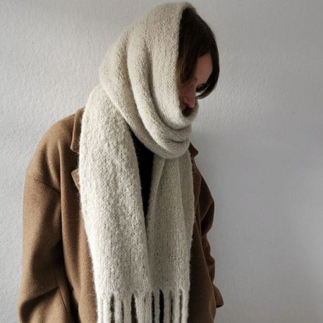 Person wearing a large, cozy beige scarf over a brown coat, stands against a plain background.